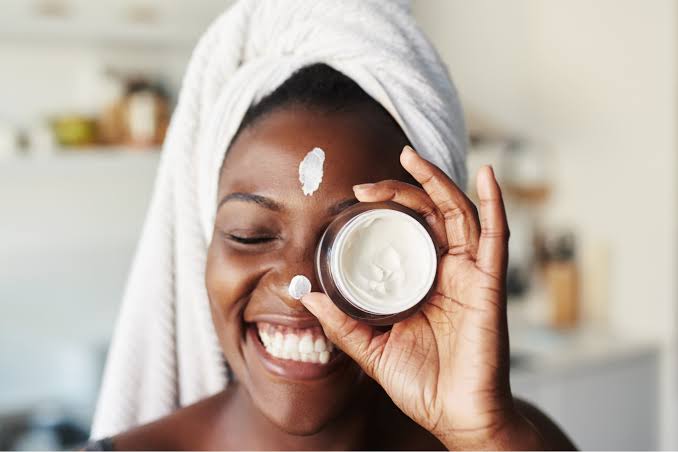 Smiling Black woman holding a skincare cream with a towel on her head